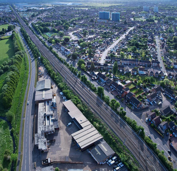 An aerial view of a residential area in Old Oak Common showing a railway line running parallel to a road and a small industrial or commercial building with a flat roof, alongside a parking area with parked cars. The neighborhood consists of closely spaced houses with gardens, streets lined with trees, and a mixture of green spaces and urban infrastructure. In the background, there are taller buildings and a broader cityscape. The scene captures the layout of the area, highlighting the proximity of residential streets to transportation routes, which is relevant to house removals and furniture transport services provided by Man with Van Old Oak Common during home relocation and packing processes.