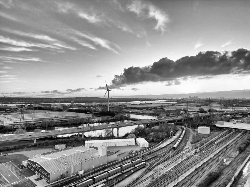 A black and white aerial photograph showing an industrial area with multiple railway tracks and a nearby warehouse building with a flat roof. Several trains are parked on the tracks, and there are large containers and storage units outside the warehouse. In the background, a road runs parallel to the railway and extends into the distance, with an overpass crossing over it. Beyond the industrial site, there are open fields, waterways, and a wind turbine with three blades visible against a cloudy sky. The scene appears to be captured during daylight hours with diffused natural lighting. This image reflects the logistical environment associated with home relocation and furniture transport, aligning with professional removals services such as those offered by Man with Van Old Oak Common.