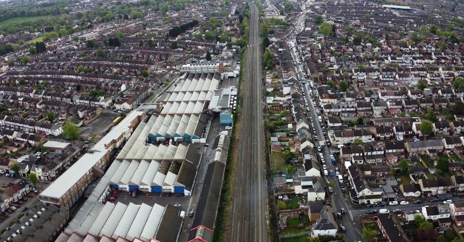 An aerial view of a residential area in Old Oak Common showing a railway line running parallel to a road and a small industrial or commercial building with a flat roof, alongside a parking area with parked cars. The neighborhood consists of closely spaced houses with gardens, streets lined with trees, and a mixture of green spaces and urban infrastructure. In the background, there are taller buildings and a broader cityscape. The scene captures the layout of the area, highlighting the proximity of residential streets to transportation routes, which is relevant to house removals and furniture transport services provided by Man with Van Old Oak Common during home relocation and packing processes.
