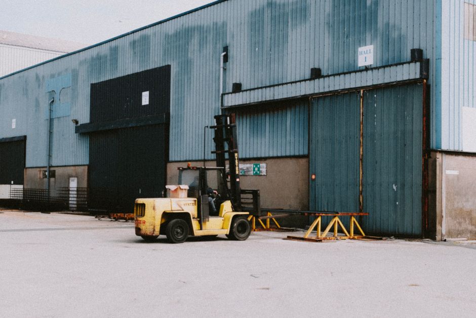 A yellow forklift truck is positioned outside a large industrial warehouse building with blue corrugated metal walls and multiple large black sliding doors. The forklift is in the process of loading or unloading heavy wooden pallets that are supported by orange metal barriers on the paved ground surface. The surrounding environment includes warehouse signage, safety instructions near the doors, and a relatively open loading bay area with minimal obstructions. The lighting appears natural, suggesting daytime conditions. This scene relates to house removals and furniture transport, illustrating the logistics involved in loading goods for relocation or storage purposes, as performed by Man with Van Old Oak Common, a professional removals company.