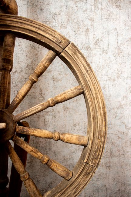 A close-up image of a large, round wooden wheel with multiple spokes, featuring a weathered, rustic appearance and a natural wood finish. The wheel is positioned vertically, leaning against a textured, light-colored wall with visible wear and subtle patterns. The wheel's rim is smooth, with some minor cracks and aging marks, indicative of its vintage or antique nature. Part of a metal hub is visible at the centre, with spokes radiating outward. This scene suggests an interior setting, possibly related to a home or storage area used for packing or preparing furniture for house removal. The detailed wooden craftsmanship could relate to furniture transport within a house relocation process, highlighting the importance of careful handling of items like antique wheels during moving services. Man with Van Old Oak Common, specialising in removals, utilises careful packing and transport methods for valuable or fragile objects such as this wheel.
