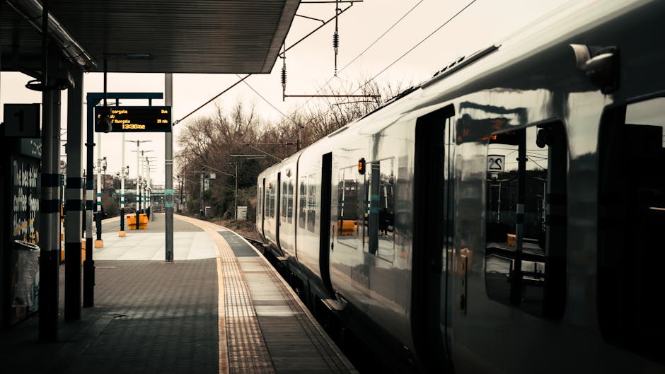 A train with a sleek, modern exterior is stationary at a railway platform. The train's reflective surface shows parts of the station environment. The platform features a textured, tactile paving strip along the edge for safety, and there are signboards displaying train times and destinations, including one indicating directions towards Old Oak Common. Overhead electrical wires extend above the train, supported by metal poles. The station's canopy provides shelter, with shadows cast on the platform. In the background, leafless trees and additional train tracks are visible, suggesting a suburban or peri-urban setting. The scene depicts a typical setting for local or regional train services, which may be used during home relocations or moving logistics for accessing properties near Old Oak Common station. Man with Van Old Oak Common specializes in removals and transportation services, including furniture and packing, supporting efficient moving processes in similar environments.