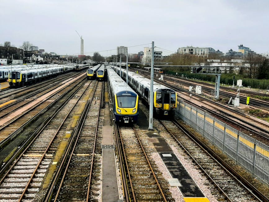 A train with a sleek, modern exterior is stationary at a railway platform. The train's reflective surface shows parts of the station environment. The platform features a textured, tactile paving strip along the edge for safety, and there are signboards displaying train times and destinations, including one indicating directions towards Old Oak Common. Overhead electrical wires extend above the train, supported by metal poles. The station's canopy provides shelter, with shadows cast on the platform. In the background, leafless trees and additional train tracks are visible, suggesting a suburban or peri-urban setting. The scene depicts a typical setting for local or regional train services, which may be used during home relocations or moving logistics for accessing properties near Old Oak Common station. Man with Van Old Oak Common specializes in removals and transportation services, including furniture and packing, supporting efficient moving processes in similar environments.