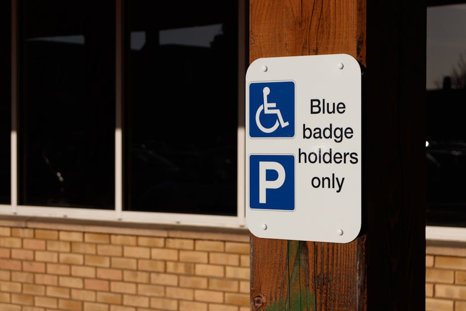 A white rectangular parking sign with a blue wheelchair symbol and text indicating 'Blue badge holders only' is mounted on a wooden post outdoors. The sign also features a blue 'P' parking symbol. Behind the sign, there is a brick building wall with large windows reflecting a dark sky, suggesting early evening or cloudy weather. The sign's positioning implies it marks designated parking spaces for disabled individuals. This scene is captured as part of a house removal or moving process, illustrating parking regulations relevant to relocation logistics near Wormwood Scrubs at Old Oak Common. The environmental lighting is subdued, emphasizing the sign's visibility and importance for accessible parking during home relocation operations, aligning with the services offered by Man with Van Old Oak Common.