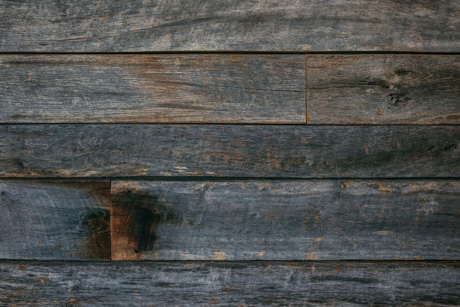 Close-up image of weathered wooden planks arranged horizontally, showing visible grain, knots, and various shades of dark brown and grey. The surface appears aged with some discoloration and minor surface imperfections. This wooden background could be part of an exterior wall or fencing. The image is well-lit, with natural lighting highlighting the texture and details of the wood. It represents a typical urban or residential setting where house removals or furniture transport may take place, aligning with the context of home relocation and moving logistics handled by Man with Van Old Oak Common. No objects, furniture, or moving equipment are visible in this shot, focusing solely on the rustic wooden surface.