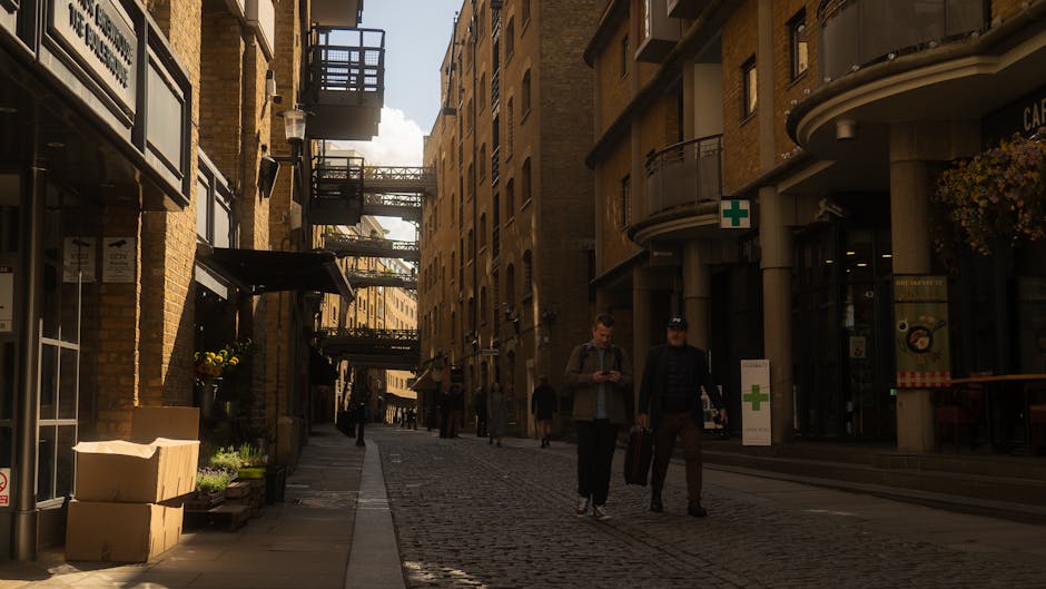 A narrow cobbled street in Old Oak Common with multi-storey brick buildings on either side, featuring external fire escape staircases and balconies. On the left side, there are cardboard boxes stacked outside a shop or café with potted plants nearby, while on the right, a building with a curved façade has a green cross sign indicating a pharmacy or health service. Several people are walking along the street, including two men in casual clothing, one with a backpack and the other carrying a briefcase, engaged in conversation. The men are near a parked van, partially visible at the bottom right corner, used for home relocation or furniture transport. The scene is illuminated by daylight, with some shadows cast on the cobblestone pavement, and additional pedestrians are visible further down the street in the distance. This setting illustrates a typical urban environment suitable for moving services such as packing and loading furniture, aligned with house removals or relocation efforts provided by Man with Van Old Oak Common.