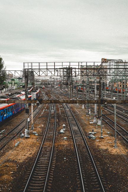 The image depicts a view of railway tracks extending into the distance, with multiple sets of steel rails and overhead electrical lines supported by metal structures. To the left, there are freight tr