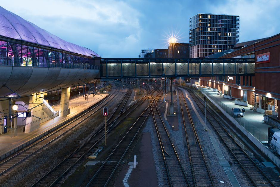 The image depicts a view of railway tracks extending into the distance, with multiple sets of steel rails and overhead electrical lines supported by metal structures. To the left, there are freight tr