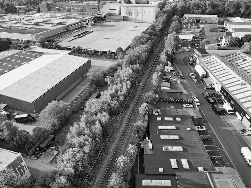 An aerial black-and-white photograph showing a commercial area with several industrial and office buildings, parking lots filled with cars, and a row of trees separating the buildings from railway tracks running vertically through the center of the image. On the left, a large warehouse with a flat roof and several smaller structures are visible, while on the right, there are additional office or retail units with designated parking spaces and vehicles parked adjacent. The railway tracks, lined by dense foliage and trees, extend from the bottom to the top of the image, suggesting the scene involves a property boundary or route used during home relocation or furniture transport. This setting, captured during daylight with clear details of building rooftops, parked vehicles, and surrounding greenery, illustrates the logistical environment typical of a house removals project coordinated by companies such as Man with Van Old Oak Common, who specialise in loading, packing, and transporting household belongings in the London area.