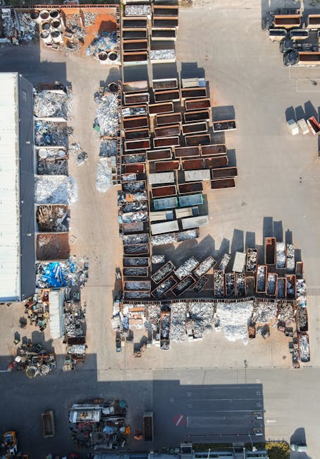 An aerial view showing numerous large metal and cardboard boxes arranged in rows on a concrete surface, with some boxes open revealing packing materials like bubble wrap and foam. To the left, there is a warehouse or storage building partially visible, and on the right, a van with open side doors with additional boxes and packing supplies nearby. In the foreground, a small area contains additional packaging materials and equipment, with a few vehicles parked along the bottom edge. The scene depicts a logistics or moving preparation area, consistent with house removals and furniture transport processes, as managed by Man with Van Old Oak Common, situated near Old Oak Common.