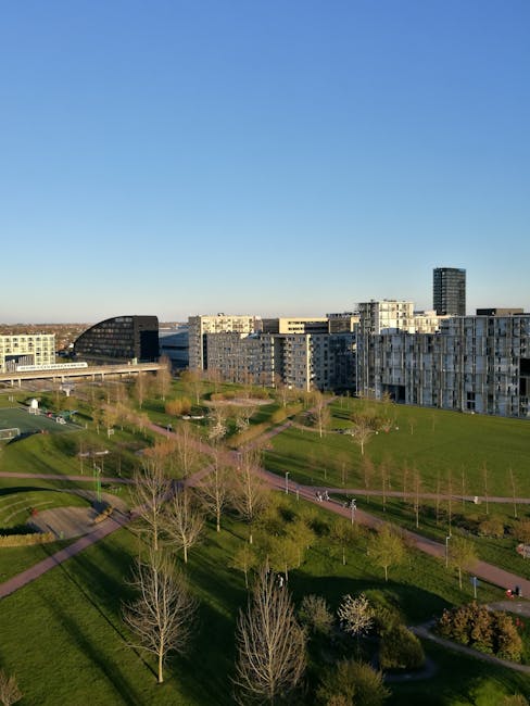 A spacious outdoor scene showing a park area with several leafless trees and walking paths on well-maintained grass, with a backdrop of modern residential and office buildings in Old Oak Common. The b